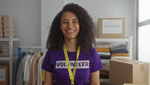 Young hispanic woman smiling in purple volunteer shirt at indoor charity center with clothing donations in the background. - Powered by Adobe