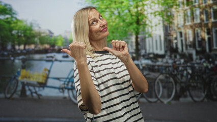 Woman with thumbs pointing to self in a striped shirt on street near canal bikes, smiling and tilting head while gesturing to chest; confidence playful.