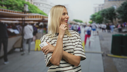 Woman holding smartphone, crossing arms and touching chin, striped shirt in street market;...
