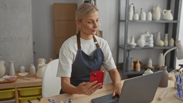 Woman potter holding red smartphone and tapping laptop in a ceramics studio while wearing denim apron and checking orders; focused productivity. - Powered by Adobe