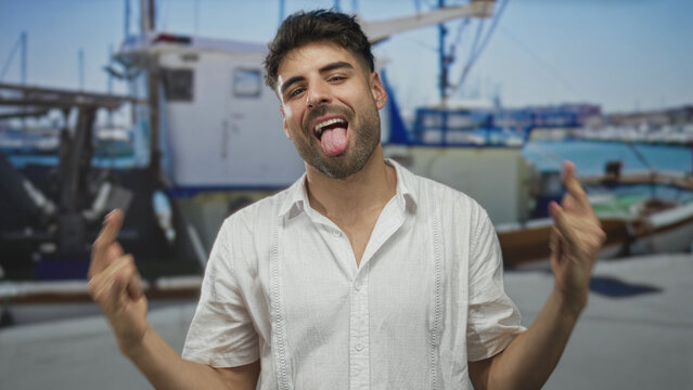 Man makes rock horns gesture with bare hands in front of boat on sunny street by seaside port; playfulness.