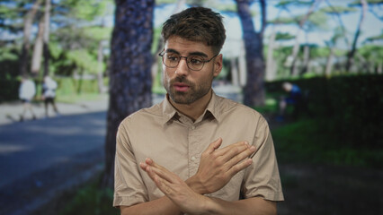 Fototapeta premium Man wearing beige shirt crossing arms in green park on sunny walkway under tall pine trees; denial frustration.