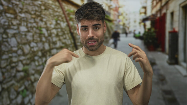 Young man finger pointing to chest in narrow street with stone wall and blurred pedestrians; uncertainty doubt hesitation confusion.