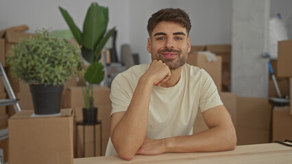 Hispanic young man rests chin on hand in newly unpacked home filled with stacked moving boxes and...
