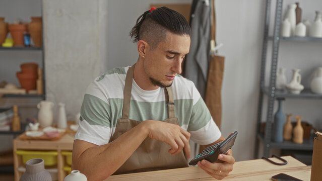 Man potter in apron tapping calculator with right hand at pottery workbench in studio; craft concentration. - Powered by Adobe