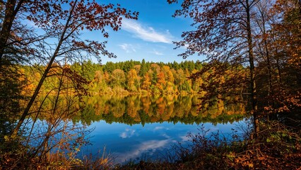 Obraz premium Autumn landscape with a lake surrounded by colorful fall trees and a blue sky with clouds.