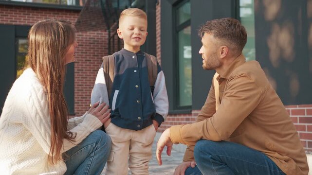 Caucasian parents crouching and talking with their son near entrance. Mother smiling while asking questions. Father listening and supporting child during conversation before heading to school.