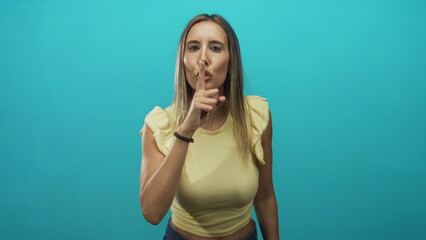 Woman with index finger to lips gesture in studio with turquoise backdrop, wearing yellow crop top and direct gaze; silence secrecy.