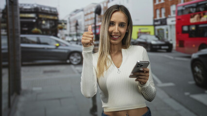 Woman with smartphone giving thumbs up on a city street while smiling and tapping screen; happiness connection.