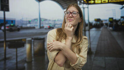 Woman wearing glasses hugs arms at airport terminal curbside under overcast sky awaiting flight;...