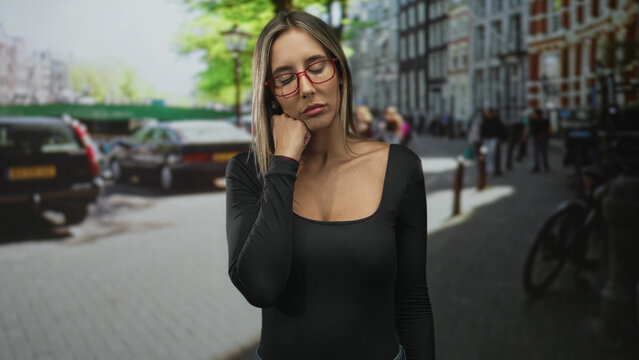 Woman resting hand on chin on city street near parked cars and bicycle rack, wearing red glasses and black long sleeve top; pensive.