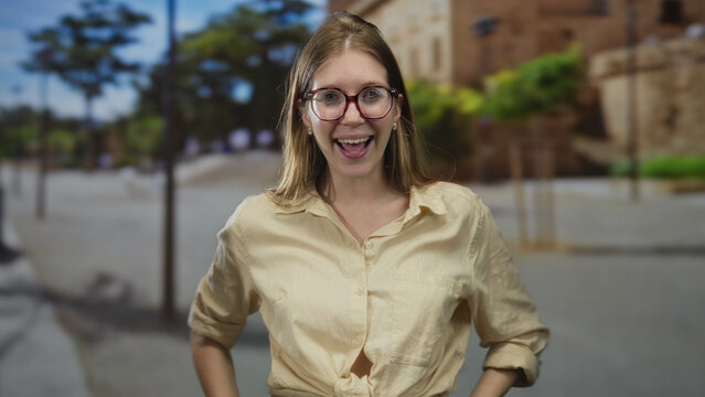 Woman with bare arms forming hand binoculars gesture on a sunlit urban street lined with buildings; exploring joy.