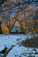 A-frame cabin in snowy forest at sunset, warm lights inside, winter retreat among leafless trees.