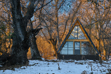 A-frame cabin in snowy forest at sunset, warm lights inside, winter retreat among leafless trees.