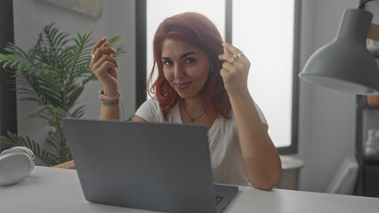 Woman at laptop touching hair and typing in studio at home desk, hands visible and wearing rings and necklace; focused productivity.