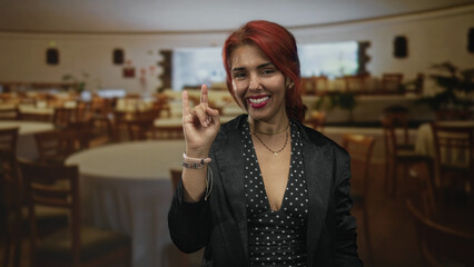 Young hispanic woman with red hair and polka dot dress, hand forming love sign inside restaurant building; friendly joy.