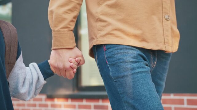 Caucasian boy walking hand in hand with parent near brick school building. Child smiling while heading away from classes. Parent guiding along pathway during relaxed after school moment.