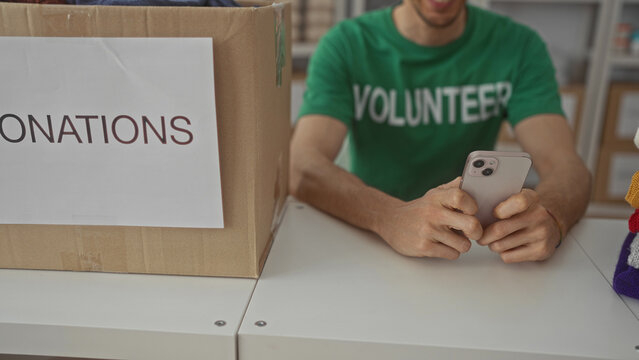 Man in green volunteer shirt holds smartphone by a pile of folded sweaters beside a cardboard donation box in a building; compassion. - Powered by Adobe