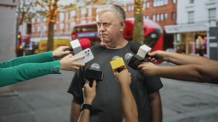 Man interviewed, microphones and smartphone held by multiple hands pointed at his face as reporters press close on a busy city street; unease scrutiny.