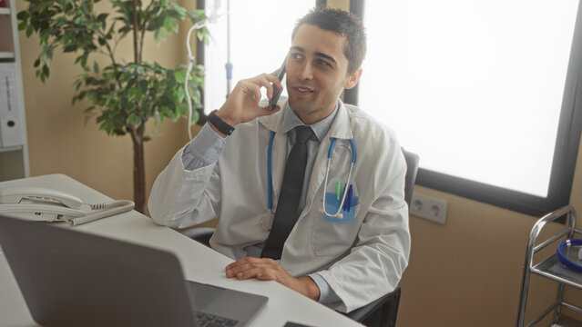 Young hispanic man doctor with stethoscope in clinic room talking on phone while sitting at desk using laptop in a well lit modern workplace suggesting professionalism