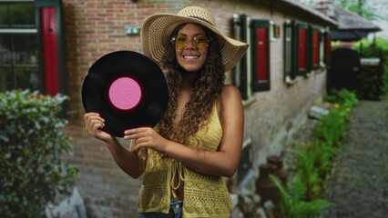 Fototapeta premium Woman holding vinyl record with both hands, wearing straw hat and yellow top, standing on a narrow street near brick wall; nostalgia summer leisure.