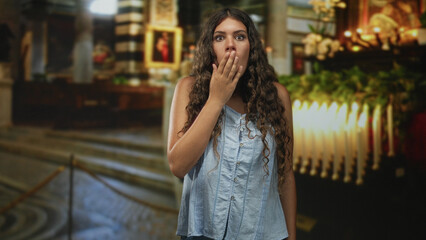 Fototapeta premium Young woman with long curly hair covering mouth with hand in church near candles, altar and ornate columns; surprise reverence.