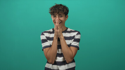 Teenage boy presses hands together covering mouth while smiling in a green studio wearing a striped shirt and visible bare forearms; playful surprise.