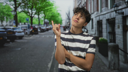 Teenage boy pointing finger upward, arms crossed, standing on a city street by parked cars and...