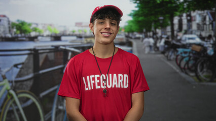 Man in red lifeguard shirt wearing whistle smiling with eyes closed on street by a canal lined with...