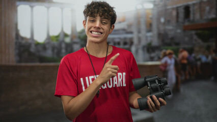 Teen lifeguard in red shirt holding binoculars and whistle points and gives thumbs up on a crowded street by stone ruins; cheerful optimism.