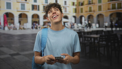 Teen boy with backpack holds smartphone and smiles while looking up on a sunlit plaza street with...