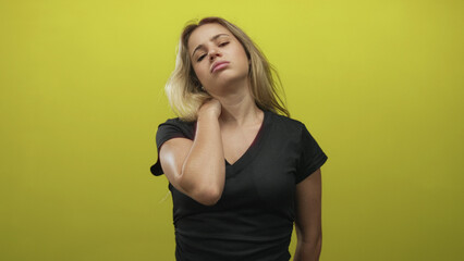Young blonde woman with hand on neck showing neckache in yellow studio, closed eyes and slumped posture, wearing black t shirt and minimal jewelry; discomfort.