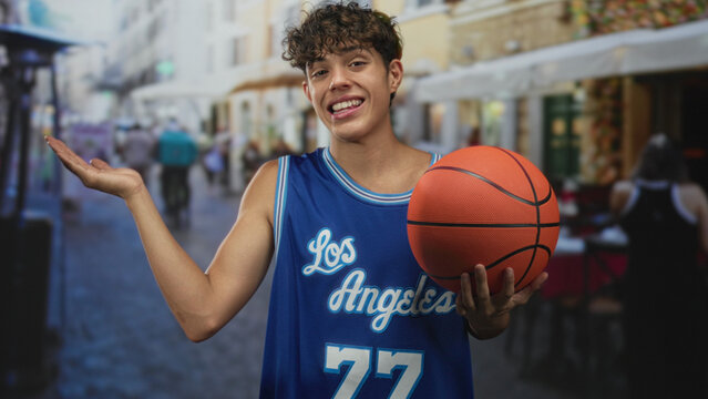 Teenage boy holding a basketball with palm up gesture on a street lined with cafes and shops; confidence.