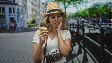 Woman holding ice cream cone with hand on cheek for toothache and vintage camera around neck on a city street; discomfort.