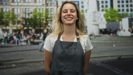 Woman with visible face smiling at camera in street plaza with buildings behind, wearing denim apron; friendly joy.