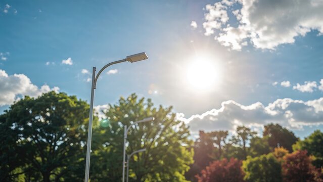 Bright sun shining above trees with streetlights under a partly cloudy sky. - Powered by Adobe