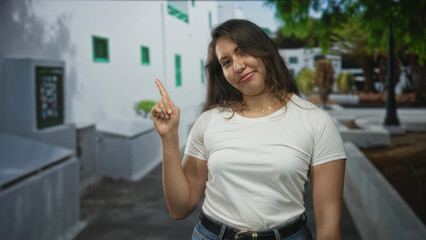 Woman points finger with bare hand at a white building on a street, casual t shirt and jeans visible, slight smile and relaxed pose; playful ease.