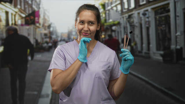 Woman holding dental tools and wearing blue gloves while smiling on a busy street with shops and pedestrians; confidence care. - Powered by Adobe