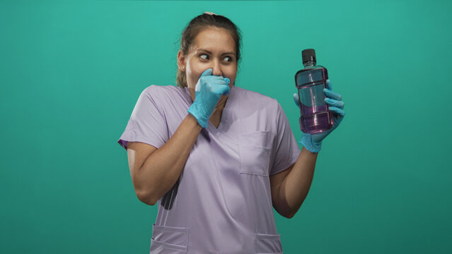 Dentist woman holding mouthwash and covering nose in studio with teal backdrop; surprise dental hygiene.