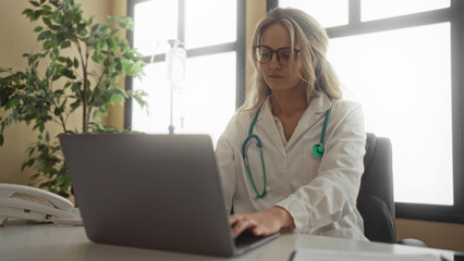 Blonde woman doctor in clinic examines laptop data wearing stethoscope, uniform, and glasses, seated in hospital office with indoor plant and medical equipment.
