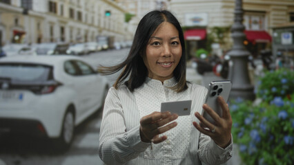 Woman holding smartphone and creditcard on city street, hands extended showing card and phone while smiling; convenience confidence.