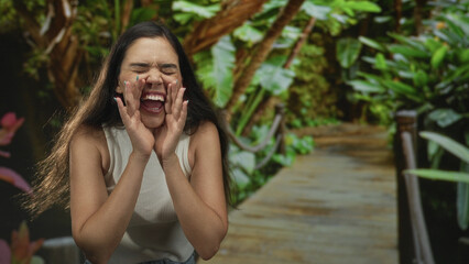 Fototapeta premium Young woman shouting with hands cupped at mouth on a sunlit wooden boardwalk in forest park surrounded by green foliage and plants; joy.
