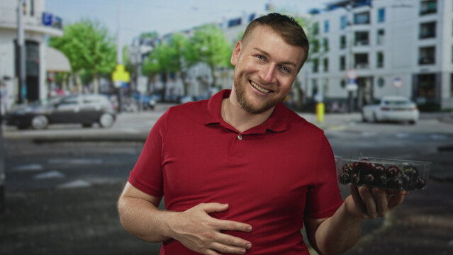 Man holding container of cherries with hand on stomach while smiling on a city street; hunger relief happiness.