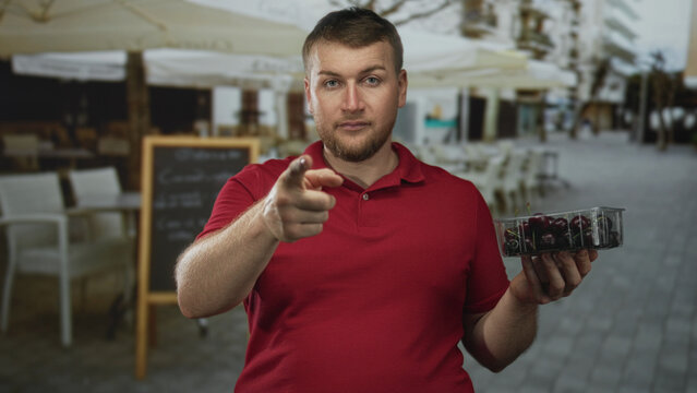 Man points finger at camera while holding a container of cherries on a restaurant terrace street; friendly offer.