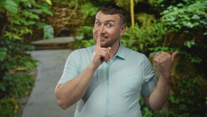 Man points thumb to forest path while wearing a light blue shirt and raising an uneasy eyebrow; confused doubt.