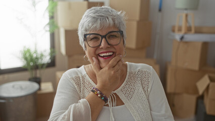 Woman with glasses covers mouth with hand while laughing among cardboard moving boxes in building; joy renewal.