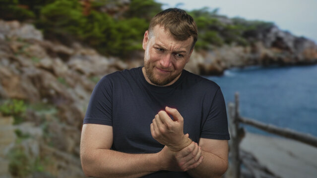 Man clutching wrist and grimacing while holding forearm on a coastal forest path by rocky cliffs and sea, wearing a dark tshirt; pain recovery.