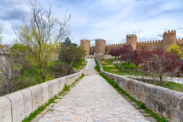 Obraz premium A cobblestone path and low stone walls lead uphill to a major gateway in the Walls of Ávila, framed by two large cylindrical stone towers under a bright, partly cloudy sky