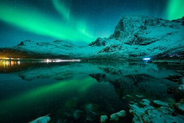 Aurora borealis over snowy mountains and calm reflective lake. Bright green Northern lights...