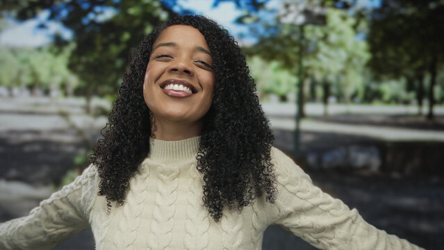 Woman tilts head upward and spreads arms amid sunlit forest wearing cream sweater by tree trunks; joy freedom connection nature.